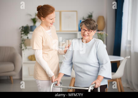 Professionelle medizinische Hausmeister in Uniform helfen Lächeln ältere Frau mit einem Walker in einem Wohnzimmer in der privaten Luxus Healthcare Clinic Stockfoto