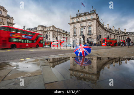 London, England - 03.15.2018: Reflexion der roten Doppeldecker Busse unterwegs am Piccadilly Circus mit britischen Regenschirm. Piccadilly Circus ist der m Stockfoto