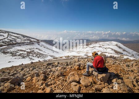 Ein junger Mann sitzt auf einem Hügel mit Blick auf die verschneiten Berge Stockfoto