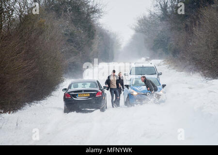 Ein Mann mit einem Spaten sein Auto aus einem Snow Drift steht in tiefem Schnee hat er die starken Winde über dem Land Straße, der wie die anderen Fahrzeuge zu kämpfen, zu graben. Stockfoto