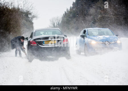 Ein Mann mit einem Spaten sein Auto aus einem Snow Drift steht in tiefem Schnee hat er die starken Winde über dem Land Straße, der wie die anderen Fahrzeuge zu kämpfen, zu graben. Stockfoto