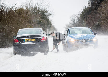 Ein Mann mit einem Spaten sein Auto aus einem Snow Drift steht in tiefem Schnee hat er die starken Winde über dem Land Straße, der wie die anderen Fahrzeuge zu kämpfen, zu graben. Stockfoto