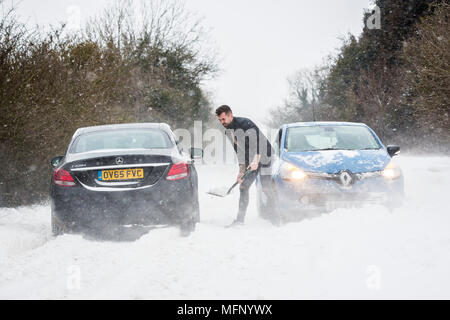 Ein Mann mit einem Spaten sein Auto aus einem Snow Drift steht in tiefem Schnee hat er die starken Winde über dem Land Straße, der wie die anderen Fahrzeuge zu kämpfen, zu graben. Stockfoto