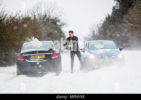 Ein Mann mit einem Spaten sein Auto aus einem Snow Drift steht in tiefem Schnee hat er die starken Winde über dem Land Straße, der wie die anderen Fahrzeuge zu kämpfen, zu graben. Stockfoto