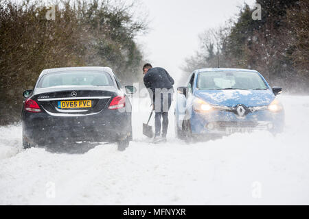 Ein Mann mit einem Spaten sein Auto aus einem Snow Drift steht in tiefem Schnee hat er die starken Winde über dem Land Straße, der wie die anderen Fahrzeuge zu kämpfen, zu graben. Stockfoto