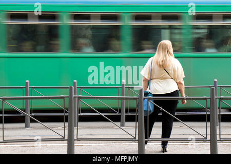 Eine blonde Frau plus Größe warten an der Straßenbahnhaltestelle in zwischen Verkehr Geländer und beobachten eine grüne Straßenbahn entgegengesetzte Richtung in Bewegungsunschärfe Stockfoto