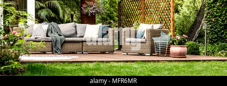Graue Sofa mit dekorativen Kissen und Körbchen mit Kissen neben Rattan Sessel stehen auf hölzernen Terrasse Stockfoto