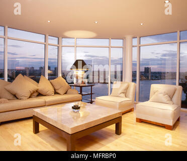 Wohnzimmer mit Holzfußboden, Couchtisch und Sofa und Stühlen vor dem Panoramafenster mit Blick auf die Stadt. Stockfoto