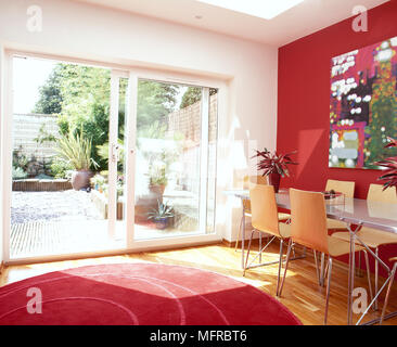 Esszimmer mit rotem Akzent Wand und Teppich, Esstisch und Stühlen, und offene Schiebetür zum Garten. Stockfoto