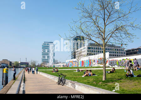 Blick auf Menschen in Riverside Park neben dem ursprünglichen Abschnitt der Berliner Mauer an der East Side Gallery in Friedrichshain, Berlin, Deutschland Stockfoto