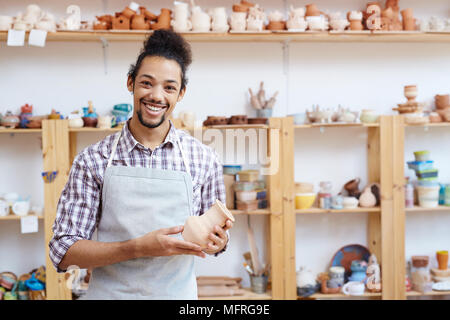 Porträt der jungen Mixed-race Mann in Schürze Holding handgefertigte Vase aus Ton in der Werkstatt und an der Kamera fröhlich lächelnd Stockfoto