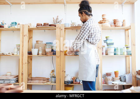 Junge afrikanische amerikanische männliche Potter in Schürze, Workshop, um beim Stehen durch Regal mit Kunsthandwerk und Ausrüstung Stockfoto