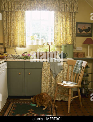 Land gelbe Küche Schränke mit grünen geblümten Vorhängen und eine Katze Essen aus einer Schüssel auf den Boden. Stockfoto