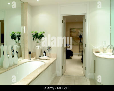 Modernes Badezimmer mit Waschbecken in gekrümmten Vanity Set, Badewanne mit Marmor umgeben, große Spiegel, und eine offene Tür zu einem angrenzenden Raum. Stockfoto