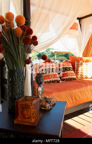 Blumen auf dem Tisch vor Tagesbett mit Baldachin und orange Decken und Kissen Stockfoto