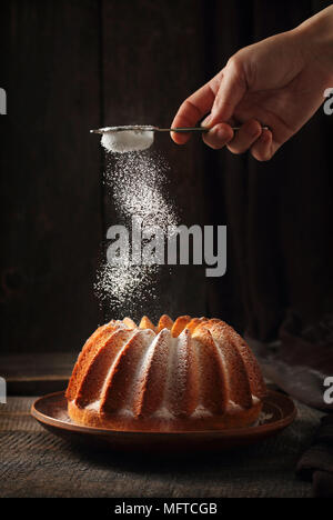 Das Streuen von orange Kuchen mit Puderzucker, dunklen Hintergrund, Licht von einem Blitz und einem Reflektor Stockfoto