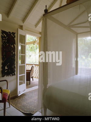 Schlafzimmer mit Himmelbett, gefliestem Boden und Holz Skulptur an der Wand mit französischen Türen zur Veranda Stockfoto