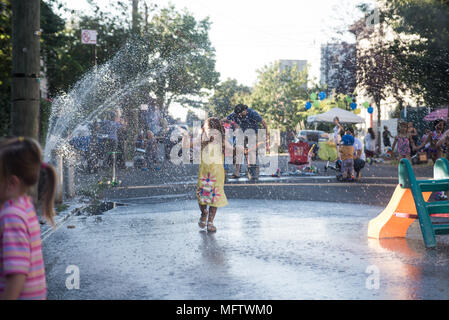 Ein siebenjähriges Mädchen in einem gelben Kleid läuft durch die Gischt eines Hydranten in einem Block Party an einem heissen Sommertag in Red Hook. Brooklyn, 2017. Stockfoto