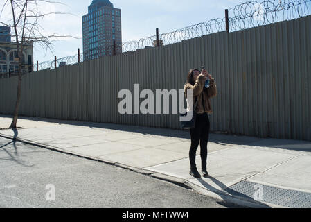 Eine junge Frau in Schwarz mit einem webpelzjacke gekleidet steht an einer Straßenecke in DUMBO und Fotografien der Himmel mit ihrem Mobiltelefon. Brooklyn, 2017 Stockfoto