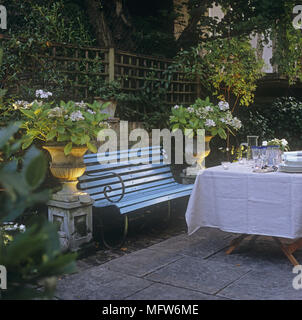 Schattig gepflasterte Terrasse mit einer Holzbank Tischdecke - gedeckten Tisch mit Geschirr und blühenden Blumen in der klassischen Stein Urnen. Stockfoto
