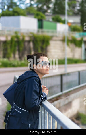 Eine junge Frau in Sonnenbrille und einem blauen Regenmantel, steht mit einem nachdenklichen Blick auf die Waterfront in der finnischen Stadt Heinola, stützte sich auf das Geländer. Stockfoto