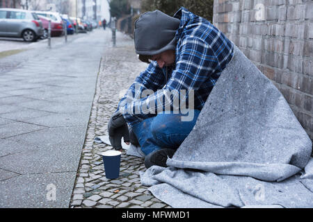 Seitenansicht eines männlichen Bettler sitzen auf der Straße in der Nähe von Einweg Becher Stockfoto