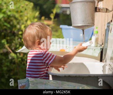 Kleines Kind, Kleinkind, 2 Jahre alt, blond, in gestreiften T-Shirt wäscht seine Hände unter Waschtisch draußen in der Landschaft Stockfoto