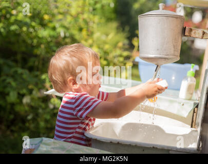 Kleiner Junge, Kleinkind, 2 Jahre alt, blond, in gestreiften T-Shirt wäscht seine Hände unter Waschtisch draußen in der Landschaft Stockfoto