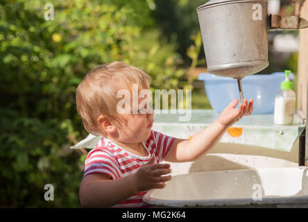 Kleines Kind, Kleinkind, 2 Jahre alt, blond, in gestreiften T-Shirt wäscht seine Hände unter Wasserspender draußen in der Landschaft Stockfoto