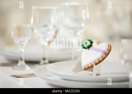 Herzförmige cookie bedeckt mit süßen Glasur, mit grünen Blumen und kleine Muster steht auf einem weißen Platten als Dekoration für festliche Hochzeitstafel in der Nähe der Wein auf dem Hintergrund Gläser eingerichtet. Stockfoto