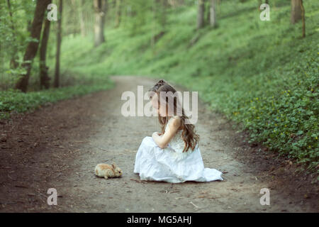 Kleines Mädchen sitzen auf der Straße in den Wald. Vor ihr sitzt ein kleiner Hase. Stockfoto