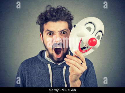 Portrait Junge verärgert wütend schreienden Mann hält ein Clown Maske auf grauen Hintergrund. Menschliche Emotionen Gefühle Stockfoto