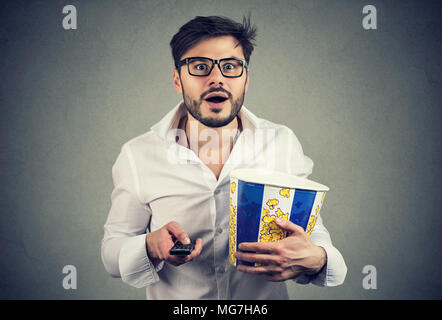 Gut aussehender bärtiger Mann in Gläsern holding Eimer Popcorn und Fernbedienung Fernsehen mit Erstaunen. Stockfoto