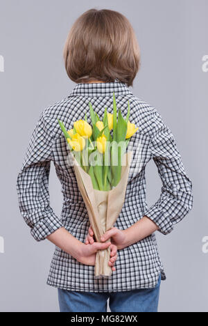 Jugendlich Mädchen verstecken Blumen hinter sich, auf grauem Hintergrund. Kind mit Strauß gelber Tulpen als Geschenk. Glückliche Mütter, Geburtstag oder Valenti Stockfoto