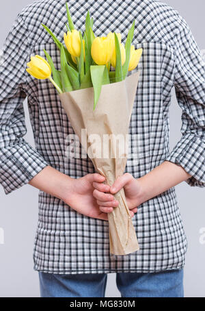 Jugendlich Mädchen verstecken Blumen hinter sich, auf grauem Hintergrund. Kind mit Strauß gelber Tulpen als Geschenk. Glückliche Mütter, Geburtstag oder Valenti Stockfoto