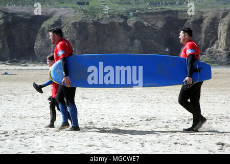 Perranporth, Cornwall, Großbritannien - 9. April 2018: Zwei Männer und ein Junge mit einem Surfbrett über den Strand in der Sonne, Rot und Blau. Stockfoto