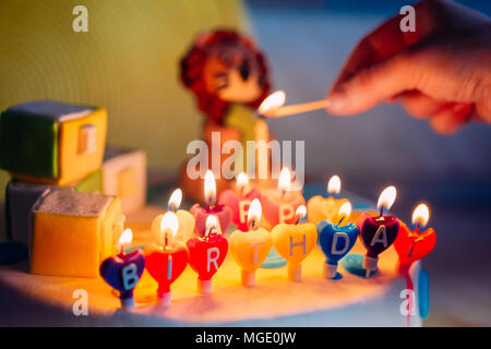 Happy Birthday In Kerzen auf bunten Kuchen geschrieben. Hand mit einem hölzernen überein, dass die Beleuchtung eine Kerze auf Kuchen. Stockfoto