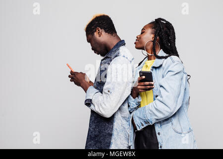 Frau versuchen Mann chatten in Handys zu sehen. Seite Portrait von afrikanischen Paar stehen Rücken an Rücken holding Handys gegen Weiße. Stockfoto