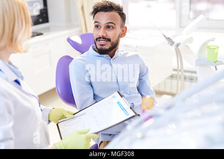 Hübscher junger Patient sitzt auf zahnmedizinischen Stuhl und im Gespräch mit Frau stomatologist, während Sie die notwendigen Hinweise in medizinische Aufzeichnung Stockfoto