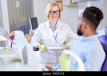 Lächelnd ziemlich stomatologist Gummihandschuhe zu tragen und weißen Mantel holding Zwischenablage in den Händen, während Fragen männlicher Patient über seine Beschwerden, Anbauteile innen Stockfoto