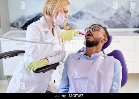 Portraitfotos von gutaussehenden jungen Mann, der zahnmedizinischen Bib sitzen auf gemütlichen Stuhl während der Zähne polieren an der Klinik Stockfoto