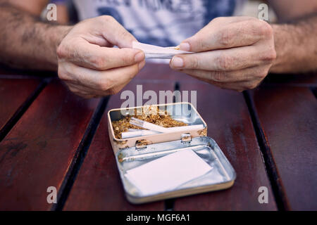 Die Hände eines Kaukasischen männlichen Rollen eine Zigarette auf einem Holztisch. Zigarettenpapier, Filter und einer Blechdose voller Tabak ist auf dem Tisch. Stockfoto