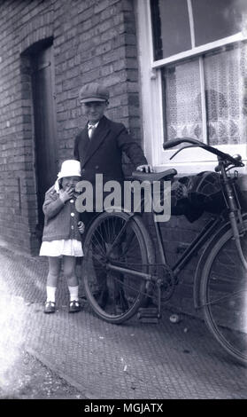 1920 s, historischen, süße kleine Mädchen steht in der Nähe gut gekleidet - ältere Bruder durch seine übergroße Fahrrad ausserhalb ihrer Reihenhaus, England, UK. Auf dem Rahmen pf das Fahrrad gibt es ein Kissen, damit er sein konnte, ihr eine Fahrt auf es irgendwo, vielleicht bis Sonntag Schule zu sehen, wie gut gekleidet sind. Stockfoto