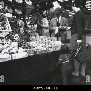 1960, London, Mutter mit Kind, ein Kleinkind an der Leine, und trägt einen Weidenkorb, Shopping im Freien an einem Obst und Gemüse marke Abschaltdruck Stockfoto