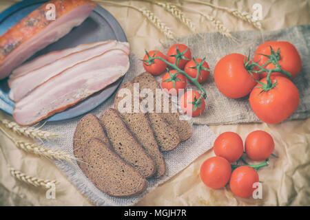In der Nähe der Scheiben Speck, Roggen, Schwarzbrot, reife rote Tomaten auf der Verpackung Handwerk Papier. Ansicht von oben. Frische Bioprodukte. Schnelle leckere Snacks. Rustikalen styl Stockfoto