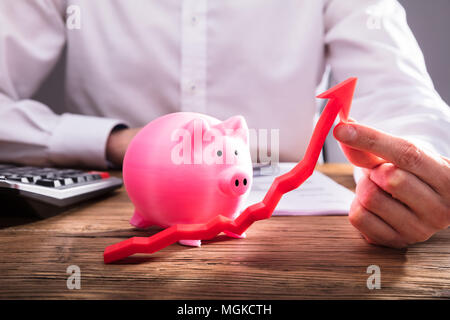 Menschliche Hand halten roten Pfeil Vor rosa Sparschwein auf hölzernen Schreibtisch Stockfoto