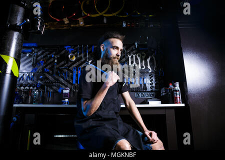 Portrait von Small Business Owner junger Mann mit Bart. Guy Fahrradmechaniker workshop Arbeiter sitzen mit Werkzeug in der Hand in einem schwarzen Kleide Stockfoto