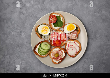 Varianten von Sandwiches mit Frischkäse, Pesto, Tomaten, Gurken, Ei, Speck, Rettiche auf grauem Hintergrund. Ansicht von oben Stockfoto