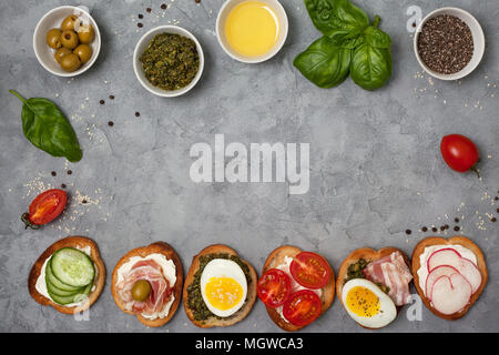 Varianten von Sandwiches mit Frischkäse, Pesto, Tomaten, Gurken, Ei, Speck, Rettiche auf grauem Hintergrund. Ansicht von oben Stockfoto