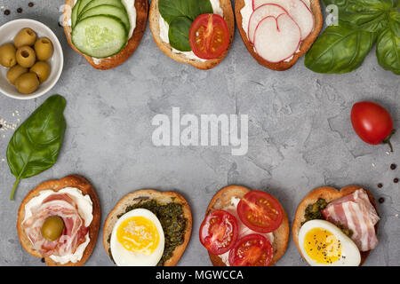 Varianten von Sandwiches mit Frischkäse, Pesto, Tomaten, Gurken, Ei, Speck, Rettiche auf grauem Hintergrund. Ansicht von oben Stockfoto